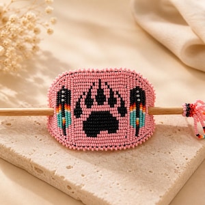 May include: A pink beaded hair slide with a black paw print and feather details. The hair accessory has a wooden stick and is presented on a textured stone surface.