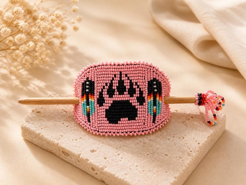 May include: A pink beaded hair slide with a black paw print and feather details. The hair accessory has a wooden stick and is presented on a textured stone surface.