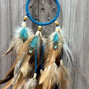 May include: A handcrafted dreamcatcher with a turquoise blue hoop and matching accents. The dreamcatcher features a woven web, wooden beads, and a collection of brown, tan, and turquoise feathers, creating a bohemian aesthetic.