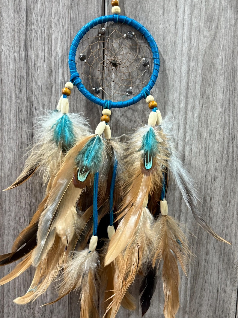 May include: A handcrafted dreamcatcher with a turquoise blue hoop and matching accents. The dreamcatcher features a woven web, wooden beads, and a collection of brown, tan, and turquoise feathers, creating a bohemian aesthetic.