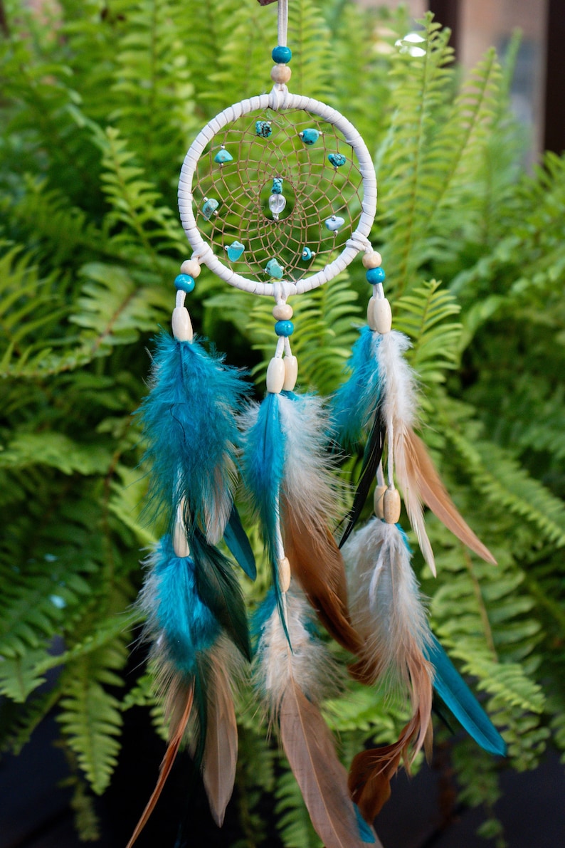 May include: A handcrafted dreamcatcher with a white hoop, adorned with turquoise stones and a woven web. Turquoise and brown feathers hang below, creating a bohemian aesthetic. The dreamcatcher is suspended against a backdrop of green foliage.