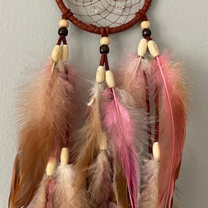 May include: A brown dreamcatcher with a woven web design and clear beads. The dreamcatcher features brown, pink, and tan feathers hanging from the bottom. Wooden beads are also included in the design.