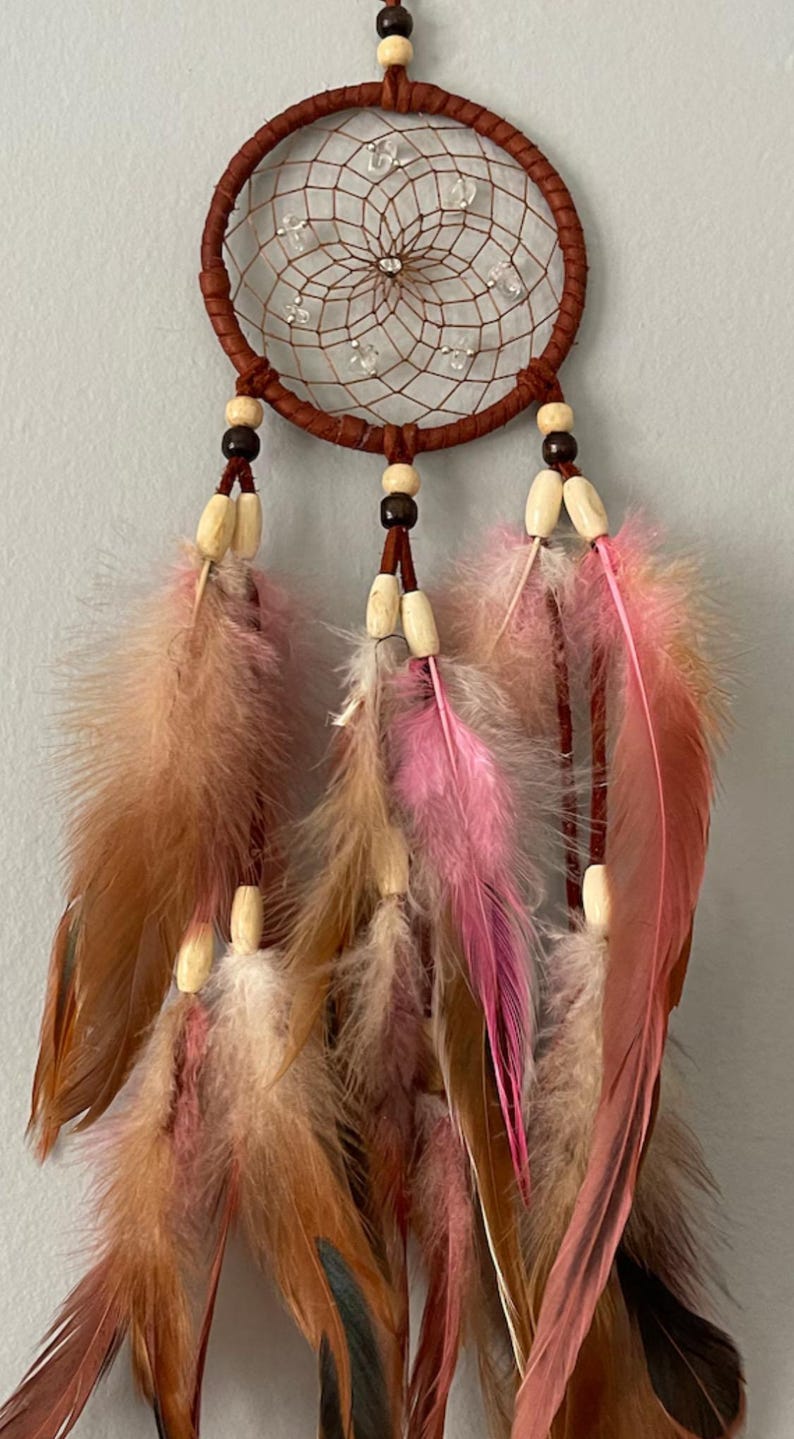 May include: A brown dreamcatcher with a woven web design and clear beads. The dreamcatcher features brown, pink, and tan feathers hanging from the bottom. Wooden beads are also included in the design.