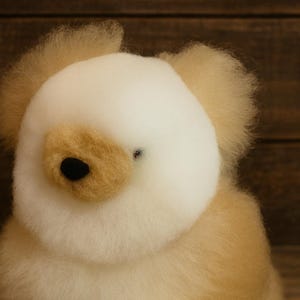 May include: A handmade, plush teddy bear with a cream-colored head and ears, and a light brown body. The bear has a black nose and a small, dark eye. The soft toy is set against a wooden background.