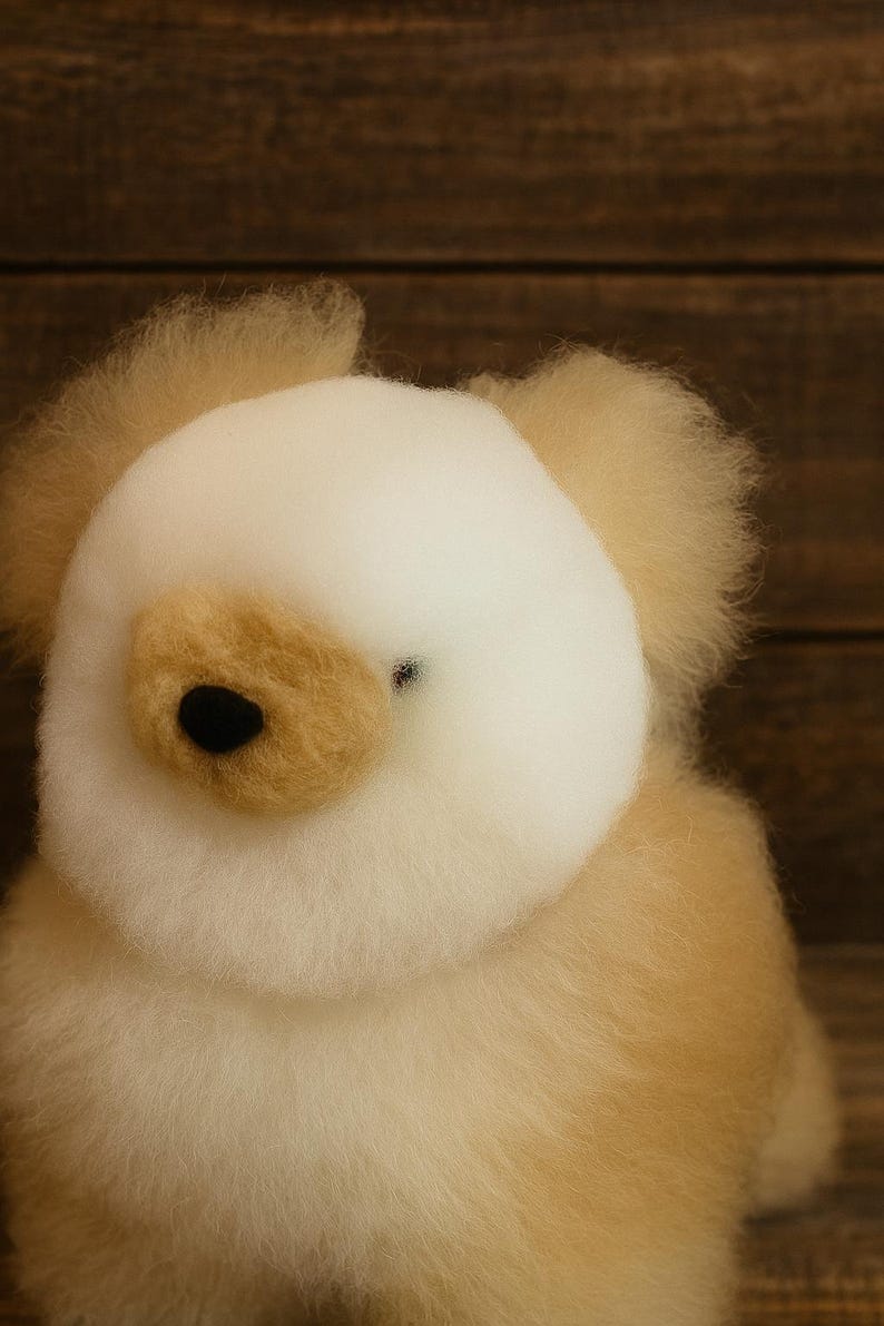 May include: A handmade, plush teddy bear with a cream-colored head and ears, and a light brown body. The bear has a black nose and a small, dark eye. The soft toy is set against a wooden background.