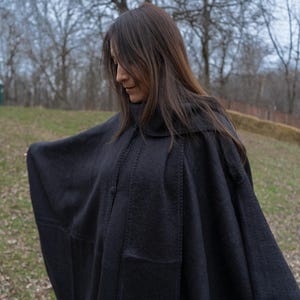 Könnte beinhalten: Ein schwarzer Wollumhang mit hohem Kragen und Knopfverschluss. Der Umhang hat ein schlichtes, elegantes Design mit sichtbaren Nahtdetails. Die Person trägt den Umhang im Freien, mit einem verschwommenen Hintergrund aus Bäumen und Gras.