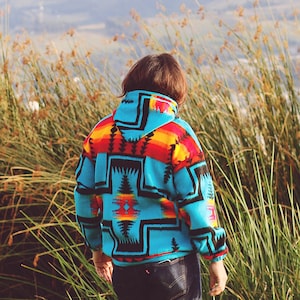May include: A turquoise fleece jacket with a Southwestern geometric pattern in red, orange, yellow, and black. The jacket has a hood. The person is wearing dark blue jeans. The person is standing outdoors near tall grass and reeds.