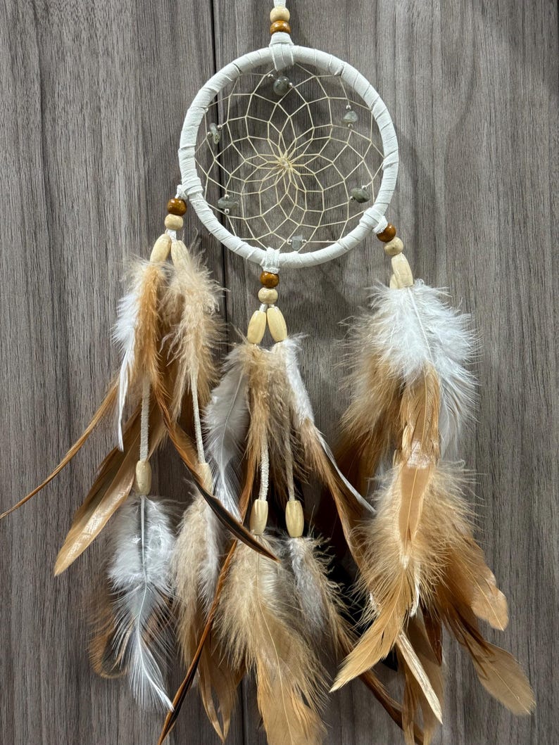 May include: A white dreamcatcher with a woven web design and hanging feathers. The feathers are a mix of brown, tan, and white, with wooden beads. The dreamcatcher is designed to hang on a wall.