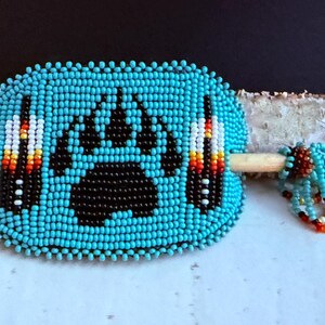 May include: A turquoise beaded hair barrette with a black bear paw print design. The barrette features a wooden stick and is adorned with colourful accents, showcasing Native American craftsmanship.