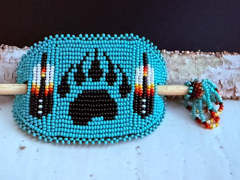 May include: A turquoise beaded hair barrette with a black bear paw print design. The barrette features a wooden stick and is adorned with colourful accents, showcasing Native American craftsmanship.