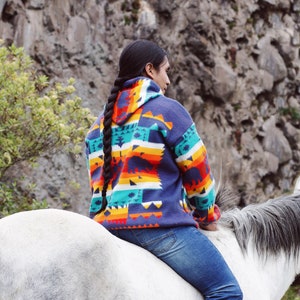 以下が含まれることがあります： 長い黒髪の三つ編みをした人が白い馬に乗っています。青いフード付きのオレンジと緑の模様のセーターと青いジーンズを着用しています。