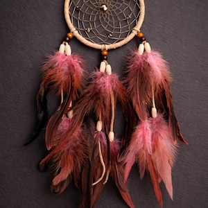 May include: A handcrafted dreamcatcher with a circular woven frame and a central web design. The dreamcatcher features hanging feathers in shades of pink and burgundy, accented with wooden beads. The background is a dark gray.
