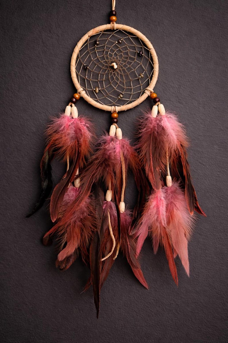 May include: A handcrafted dreamcatcher with a circular woven frame and a central web design. The dreamcatcher features hanging feathers in shades of pink and burgundy, accented with wooden beads. The background is a dark gray.