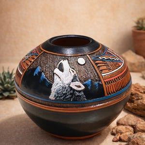 May include: A hand-painted, spherical ceramic vase with a howling wolf design. The vase features a black base and rim, with detailed artwork in shades of brown, orange, blue, and white. The wolf is depicted against a night sky with a full moon.