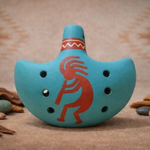 May include: A turquoise ceramic ocarina with a Kokopelli design in red. The wind instrument has six finger holes and a decorative zigzag pattern. The ocarina is displayed on a textured surface with stones and a patterned background.