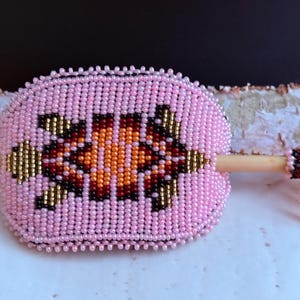 May include: Pink beaded hair barrette with a central geometric design in orange, brown, and gold. The barrette features a wooden stick and a beaded tassel. This handcrafted accessory adds a touch of colour.