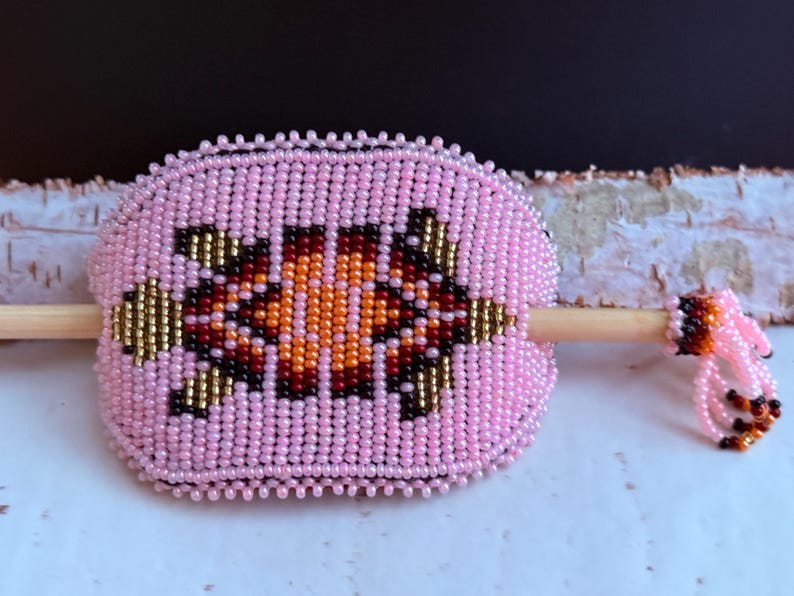 May include: Pink beaded hair barrette with a central geometric design in orange, brown, and gold. The barrette features a wooden stick and a beaded tassel. This handcrafted accessory adds a touch of colour.