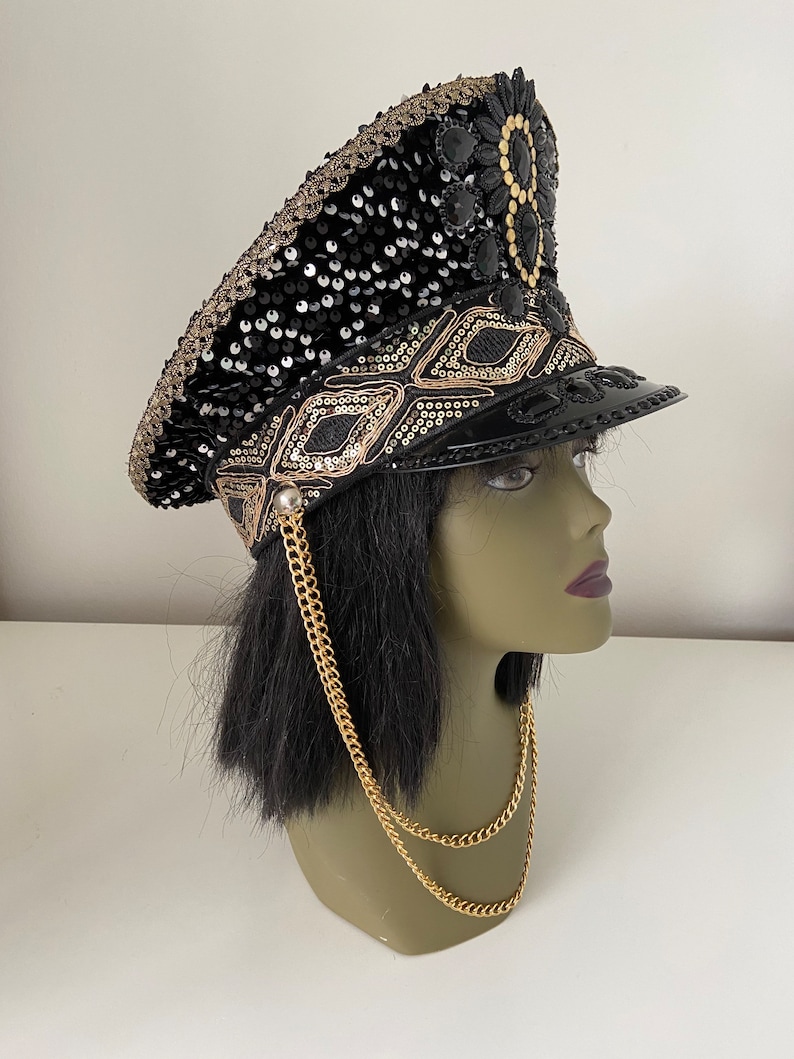 K&ouml;nnte beinhalten: Ein schwarzer Milit&auml;rhut aus Pailletten und Perlen mit einem goldenen Kettendetail. Der Hut hat ein schwarzes Schild und ein gold-schwarzes Band mit Muster.