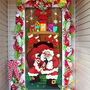 May include: A Christmas door decoration featuring a cartoon Santa Claus carrying a bag of gifts, standing in front of a fireplace with stockings hanging from it. The door is decorated with a wreath and garland.