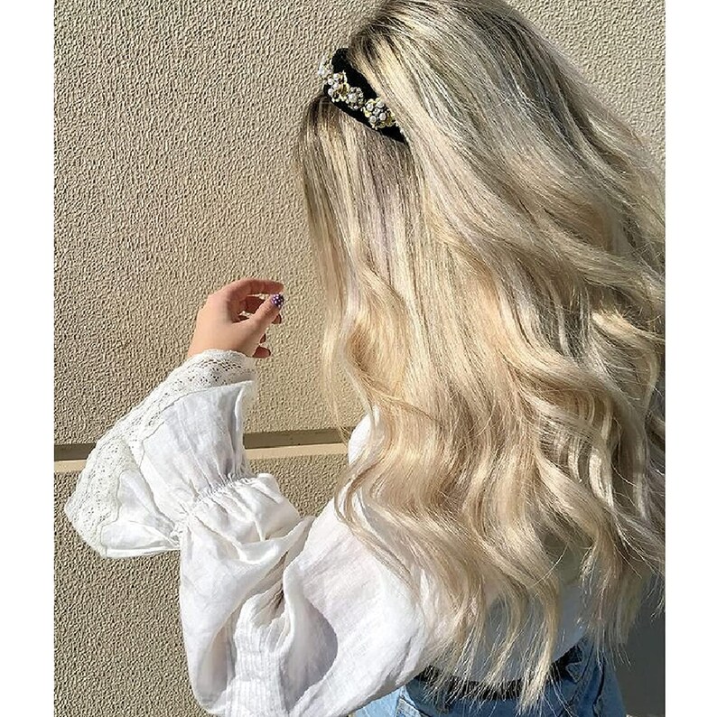 May include: A woman with long, wavy, light blonde hair wearing a white blouse with lace detailing on the sleeves. She is wearing a black headband with pearl embellishments.