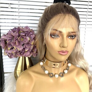 May include: A blonde wig with a light brown root, styled with a side part and bangs. The wig is on a mannequin head with a gold choker necklace with white pearls.
