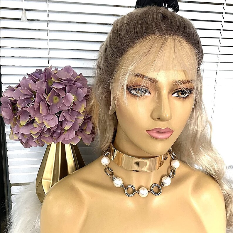 May include: A blonde wig with a light brown root, styled with a side part and bangs. The wig is on a mannequin head with a gold choker necklace with white pearls.