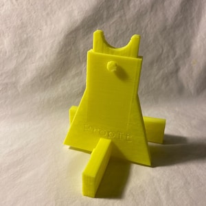 以下が含まれることがあります： 「Prop It」の文字が入った、鮮やかな黄色の3Dプリントスタンド。スタンドは、湾曲した上部と2つの支持脚を持つユニークな形状をしています。アイテムを保持するように設計されています。