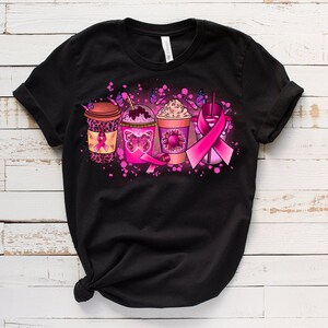 May include: Black t-shirt with a pink ribbon design featuring four coffee cups with pink and brown accents. The cups are decorated with butterflies and pink ribbons.