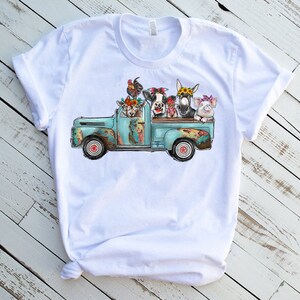 May include: A white t-shirt featuring a graphic of a blue vintage pickup truck carrying a cow, pig, donkey, goat, and rooster. The animals are wearing flower crowns.