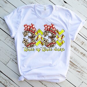 Peut inclure: T-shirt blanc avec un graphique de deux num&eacute;ros de softball "3" avec un imprim&eacute; l&eacute;opard et des n&oelig;uds rouges. Le texte "Three up Three down" est sous les num&eacute;ros.