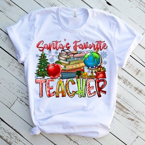 Puede incluir: Una camiseta blanca de manga corta con un estampado gr&aacute;fico que incluye el texto Santa's Favorite TEACHER. El dise&ntilde;o muestra una pila de libros, un globo terr&aacute;queo, una manzana roja, un &aacute;rbol de Navidad y luces decorativas.