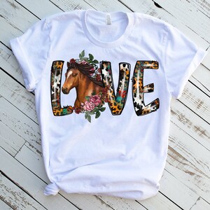 May include: White t-shirt with a graphic of the word "LOVE" in a colorful, animal print design. The letter "L" features a brown horse head with floral accents.