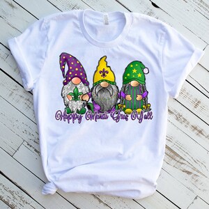 May include: White t-shirt with a graphic of three gnomes wearing Mardi Gras colors and hats. The text "Happy Mardi Gras Y'all" is printed below the gnomes.