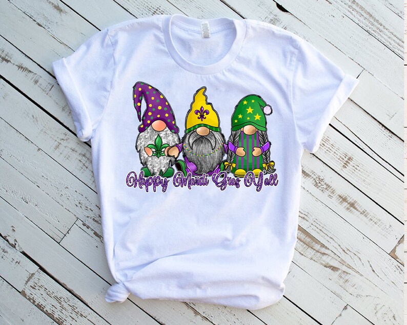 May include: White t-shirt with a graphic of three gnomes wearing Mardi Gras colors and hats. The text "Happy Mardi Gras Y'all" is printed below the gnomes.
