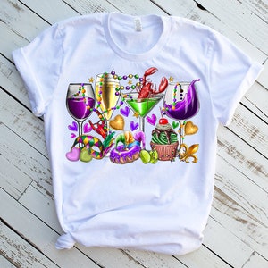 May include: White t-shirt with a colorful Mardi Gras design featuring glasses of wine, a martini, a cupcake, and beads.