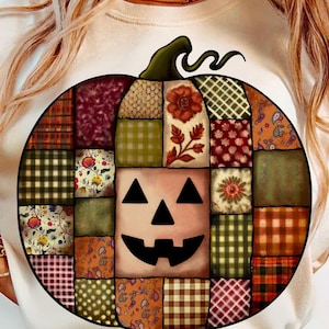 May include: A white sweatshirt featuring a large, circular pumpkin design. The pumpkin is made of patchwork squares in various patterns and colors, with a carved face in the center. The design has a green stem and curly vine.