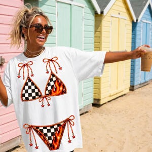 K&ouml;nnte beinhalten: Wei&szlig;es T-Shirt mit Bikini-Design. Das Bikini-Oberteil und -Unterteil haben ein Schachbrettmuster mit Flammen. Rote Schleifen und Sternakzente zieren das Bikini-Oberteil. Das Shirt ist ein l&auml;ssiger, &uuml;bergro&szlig;er Stil.