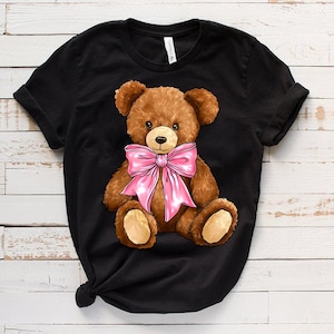 May include: Black t-shirt with a brown teddy bear wearing a pink bow. The teddy bear is sitting with its legs crossed and has a friendly expression.