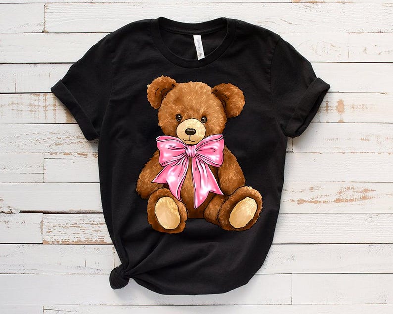 May include: Black t-shirt with a brown teddy bear wearing a pink bow. The teddy bear is sitting with its legs crossed and has a friendly expression.