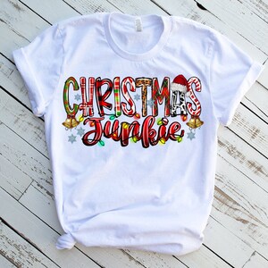 May include: White t-shirt with the text "CHRISTMAS JUNKIE" in a colorful, festive design. The text incorporates patterns like plaid, leopard print, and candy canes, with Christmas lights and bells. A Santa hat is also included.