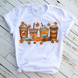 K&ouml;nnte beinhalten: Wei&szlig;es T-Shirt mit einem Aufdruck von vier Herbst-Kaffeegetr&auml;nken mit K&uuml;rbissen, Bl&auml;ttern und Eicheln.