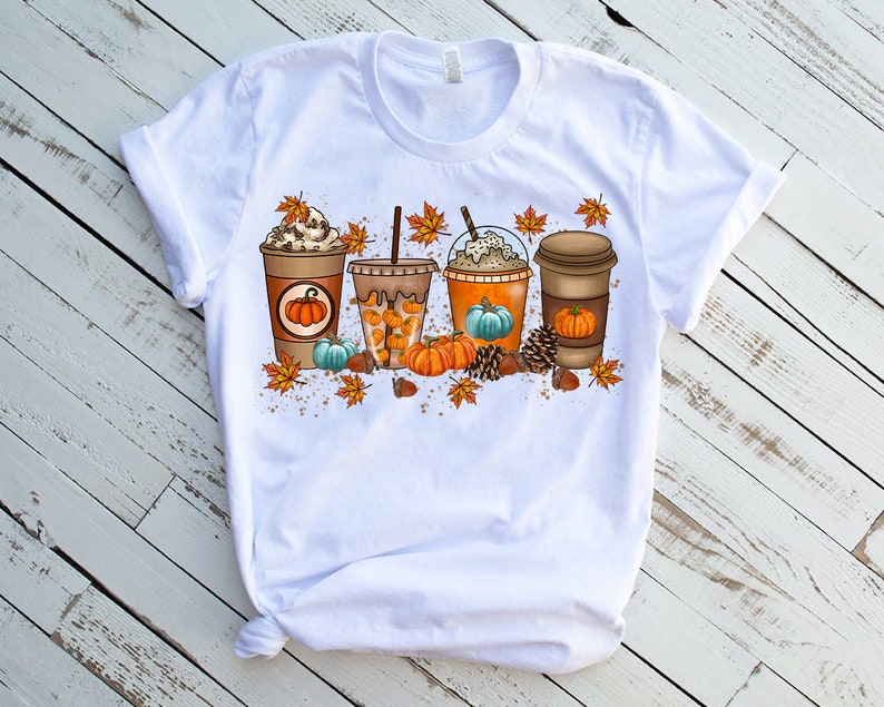 K&ouml;nnte beinhalten: Wei&szlig;es T-Shirt mit einem Aufdruck von vier Herbst-Kaffeegetr&auml;nken mit K&uuml;rbissen, Bl&auml;ttern und Eicheln.