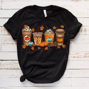 K&ouml;nnte beinhalten: Schwarzes T-Shirt mit einem Aufdruck von vier Herbst-Kaffeegetr&auml;nken mit K&uuml;rbissen und Bl&auml;ttern.