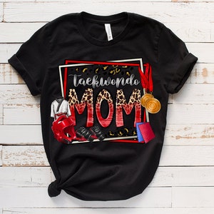 May include: Black t-shirt with the words "Taekwondo MOM" in a red and leopard print design. The shirt features illustrations of Taekwondo gear, including a uniform, headgear, gloves, and medals. The design is framed with a red and white border.