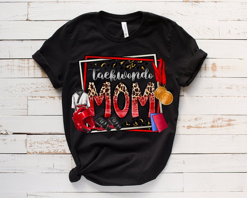 May include: Black t-shirt with the words "Taekwondo MOM" in a red and leopard print design. The shirt features illustrations of Taekwondo gear, including a uniform, headgear, gloves, and medals. The design is framed with a red and white border.