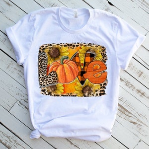 May include: White t-shirt featuring a graphic design of the word "LOVE" in orange and brown with a pumpkin and sunflowers. The design has a leopard print background.