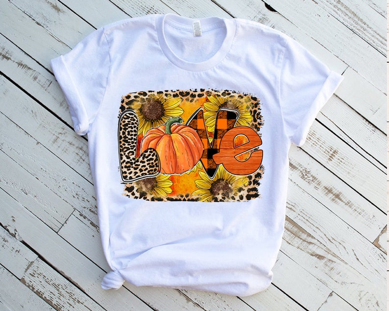 May include: White t-shirt featuring a graphic design of the word "LOVE" in orange and brown with a pumpkin and sunflowers. The design has a leopard print background.