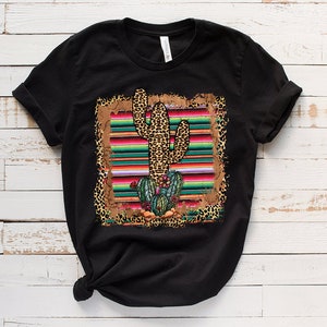 May include: Black t-shirt with a graphic print of a cactus in a colorful striped serape with leopard print accents.