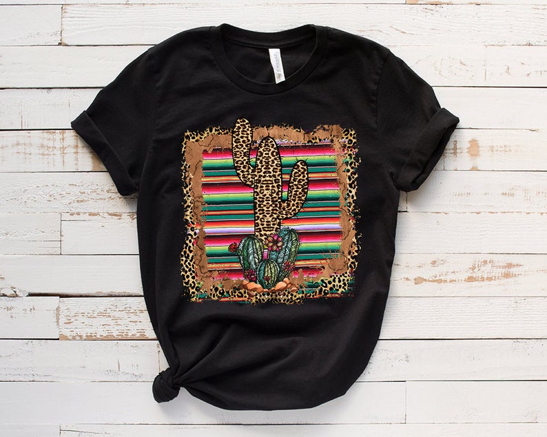May include: Black t-shirt with a graphic print of a cactus in a colorful striped serape with leopard print accents.