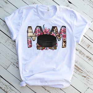 May include: White t-shirt with a floral design that spells out "MOM" with a hockey puck in the center.